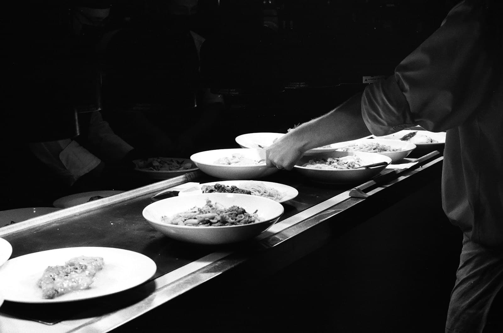A cook's arm reaching over a row of plated pasta dishes on a dark kitchen pass, in black and white.